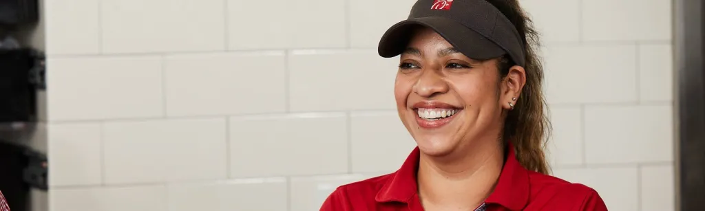 Miembro del equipo de Chick-fil-A sonriendo con una camisa roja y una visera frente a una pared de azulejos blancos.