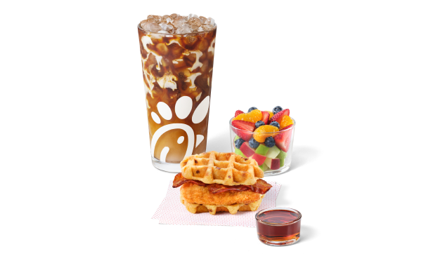 Chicken & Waffles Breakfast Sandwich, servido sobre servilletas, con una porción de syrup clásico al lado, acompañado de un Café Latte y una Ensalada de Frutas